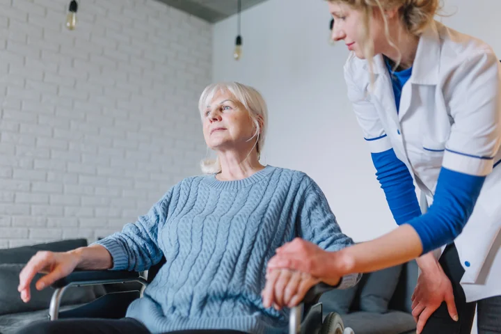 Starsza kobieta na wózku inwalidzkim, pielęgniarka trzyma jej dłoń.