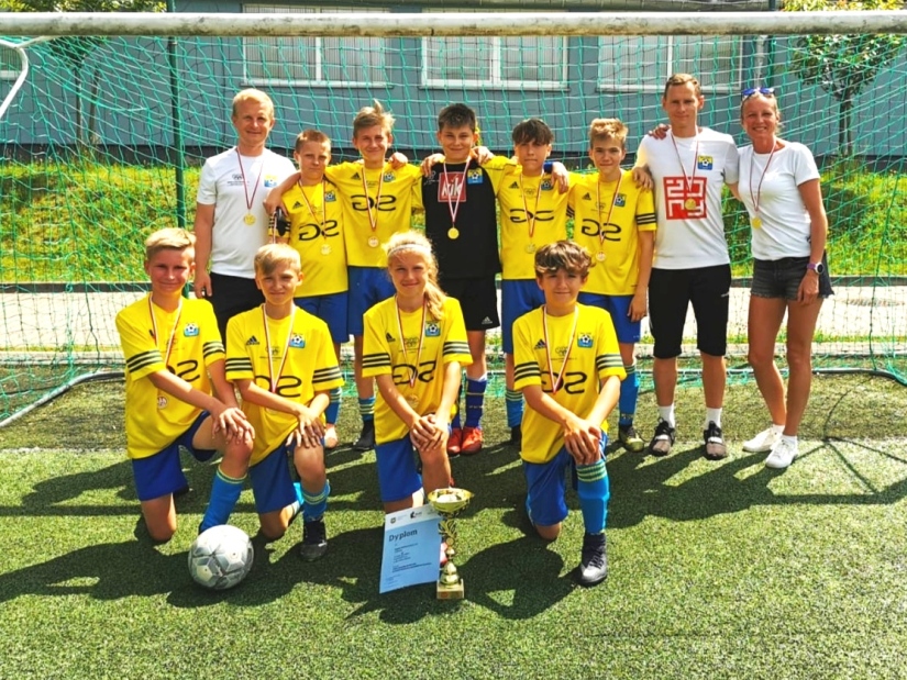 Na zdjęciu zwycięska drużyna SP 13 w żółtych koszulkach i niebieskich spodenkach wraz z trenerami.  Przed drużyną puchar, dyplom oraz piłka. 