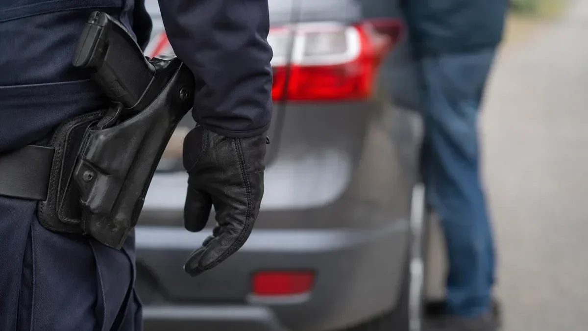 Fragment munduru policjanta z kaburą i rękawiczką, w tle tył samochodu osobowego.