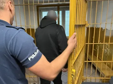 Funkcionariusz policji w niebieskim mundurze prowadzi mężczyznę w czarnej bluzie z kapturem przez żółte, metalowe drzwi więzienne.