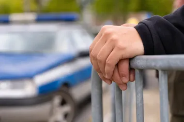 Dłonie osoby w ciemnym ubraniu oparte na metalowej barierce, w tle rozmyty policyjny radiowóz.