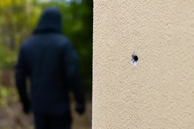 Rozmyta postać w czarnej kurtce z kapturem stoi przy ścianie z widocznym otworem po kuli.